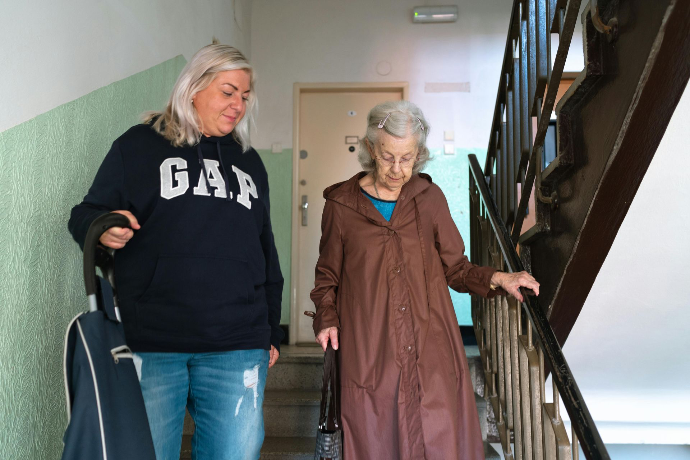Photo représentant une accompagnante soutenant une personne âgée lors de la descente d’escaliers dans un immeuble. La senior s’appuie sur la rampe pendant que l’intervenante reste à ses côtés, symbolisant l’aide à la mobilité, l’accompagnement des seniors, le maintien à domicile et la présence rassurante au quotidien. Cette image illustre les services d’aide aux personnes âgées, le transport accompagné et l’assistance personnalisée.