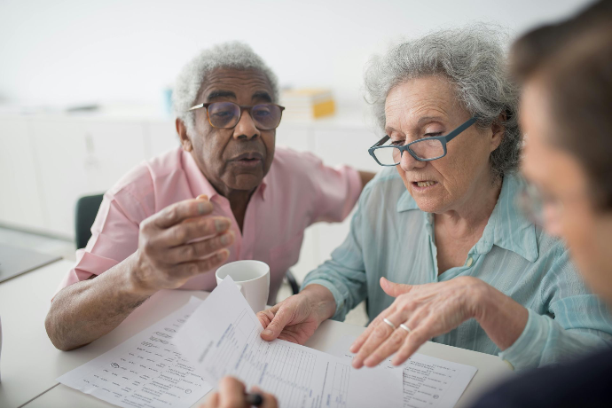 Photo représentant un couple de personnes âgées échangeant avec un accompagnant autour de documents administratifs. La scène illustre l’aide aux démarches, l’accompagnement social, la compréhension de formulaires et le soutien personnalisé aux seniors. Cette image met en avant le rôle de l’assistance administrative, le lien humain et l’accompagnement bienveillant pour faciliter les démarches du quotidien.